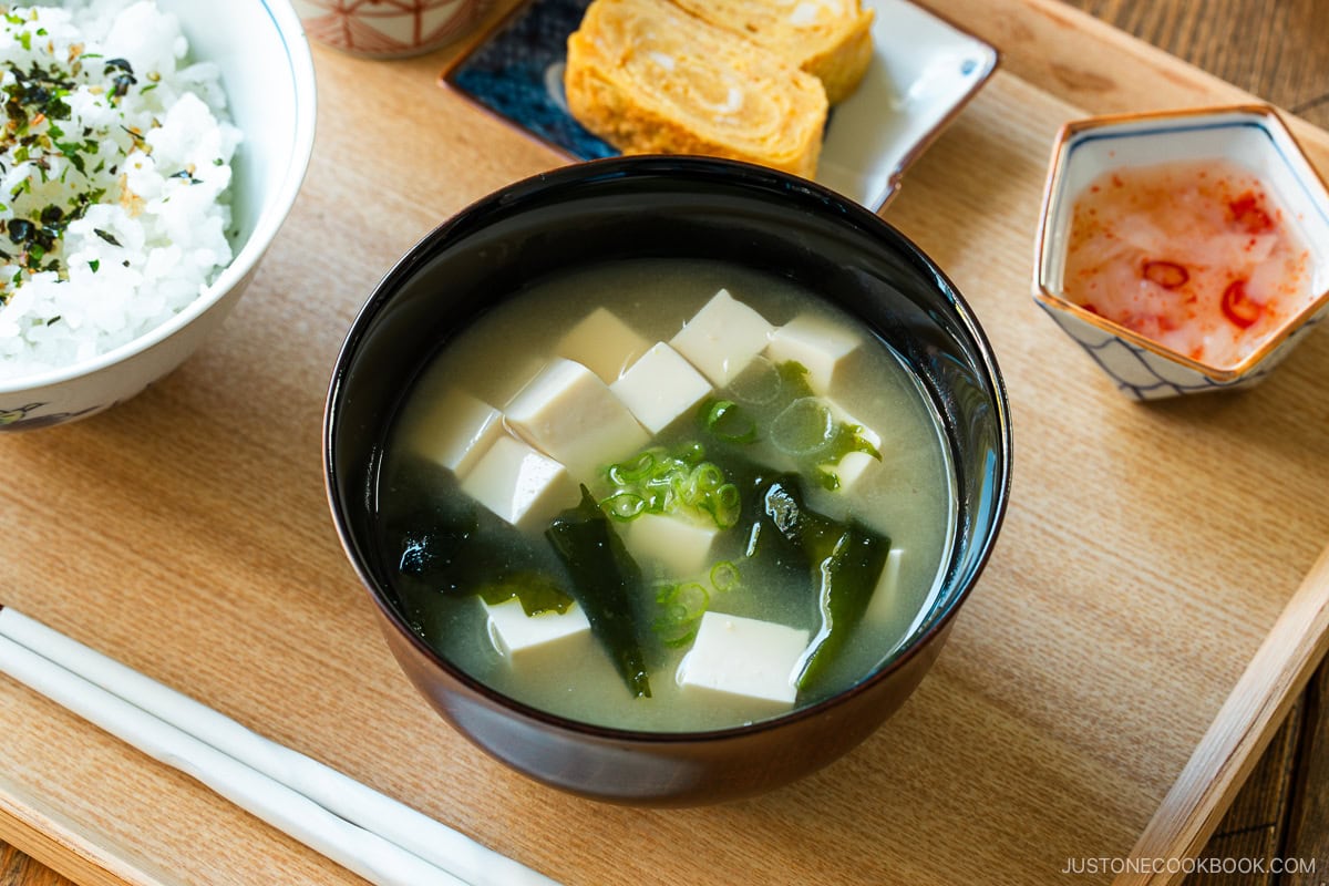 A light-colored wooden tray containing a bowl of miso soup with soft tofu, wakame seaweed, and thinly sliced green onions, along with a bowl of steamed rice, a small plate of tamagoyaki, and a smalll side dish.