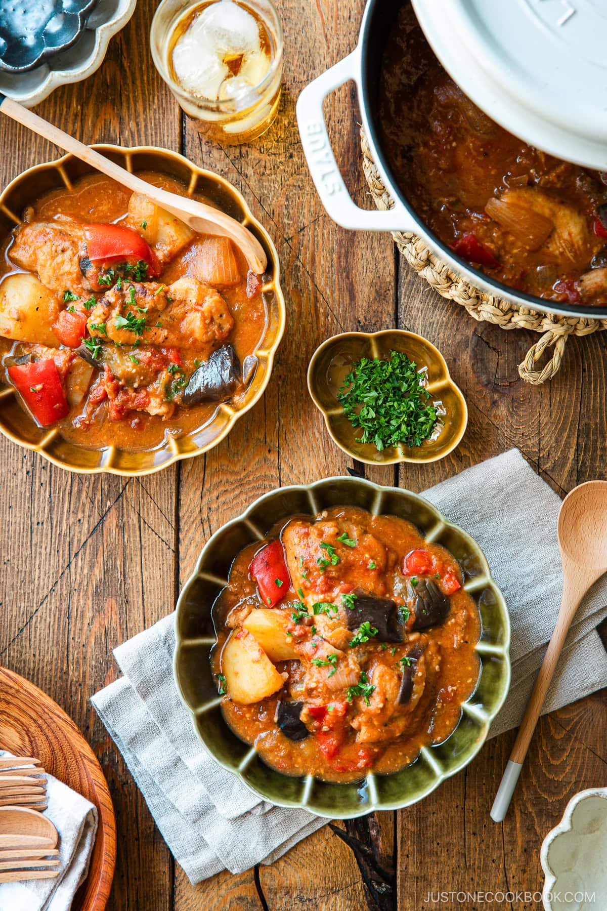 Fluted bowls containing Miso Tomato Chicken Stew garnished with chopped parsley on top, served with a glass of tea, a small dish of parsley, and a pot of the stew on the wooden table.