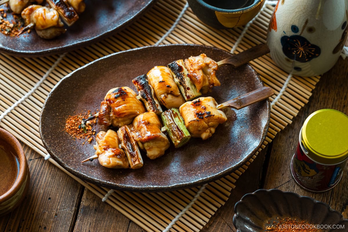 Oval rustic plate containing yakitori, Japanese grilled chicken and scallion skewers, coated with savory yakitori sauce and served with shichimi togarashi on the side.
