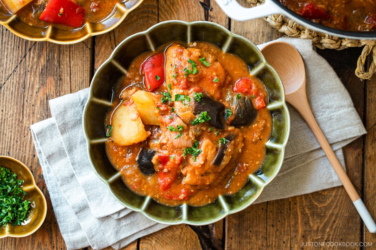 Fluted bowls containing Miso Tomato Chicken Stew garnished with chopped parsley on top, served with a glass of tea, a small dish of parsley, and a pot of the stew on the wooden table.