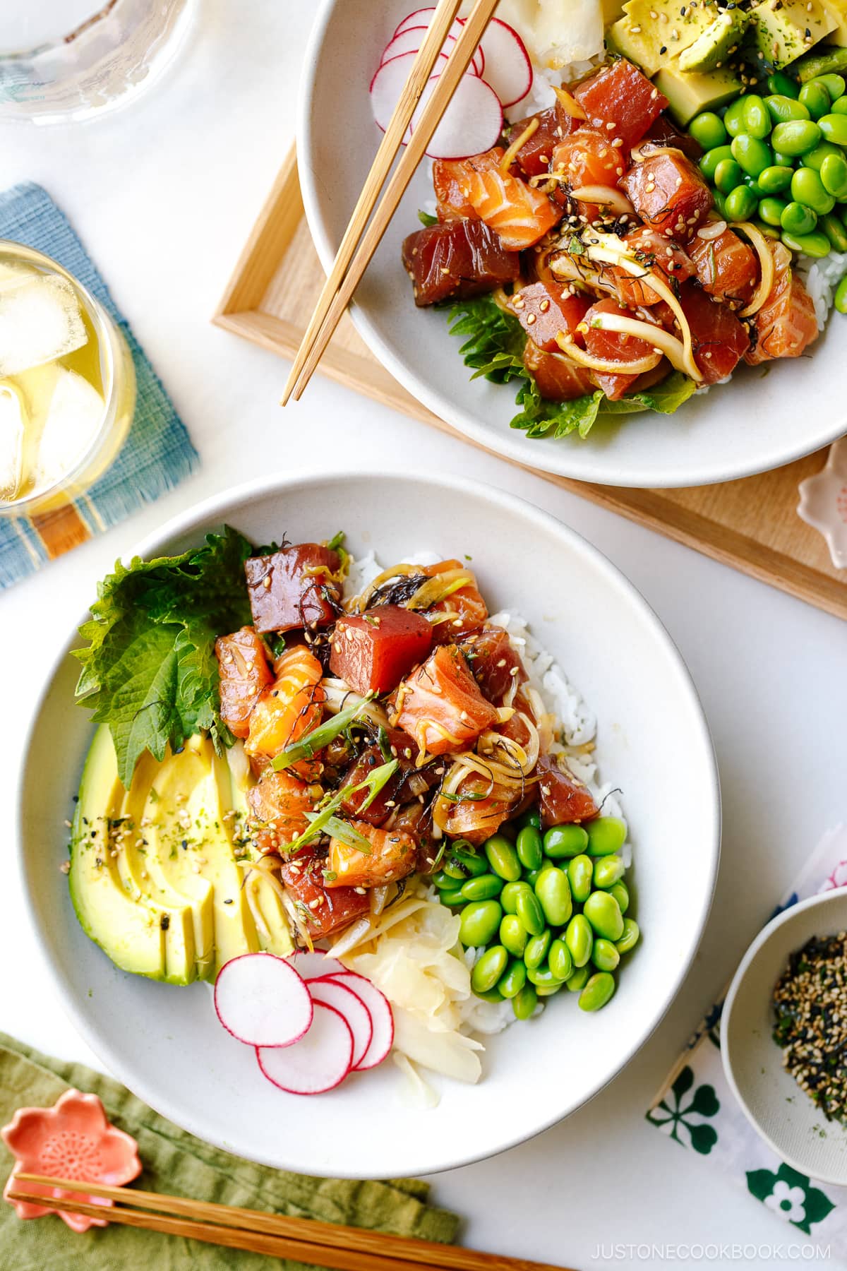 A bowl containing ahi tuna and salmon poke along with edamame, avocado, sushi ginger, and radish over bed of steamed rice.