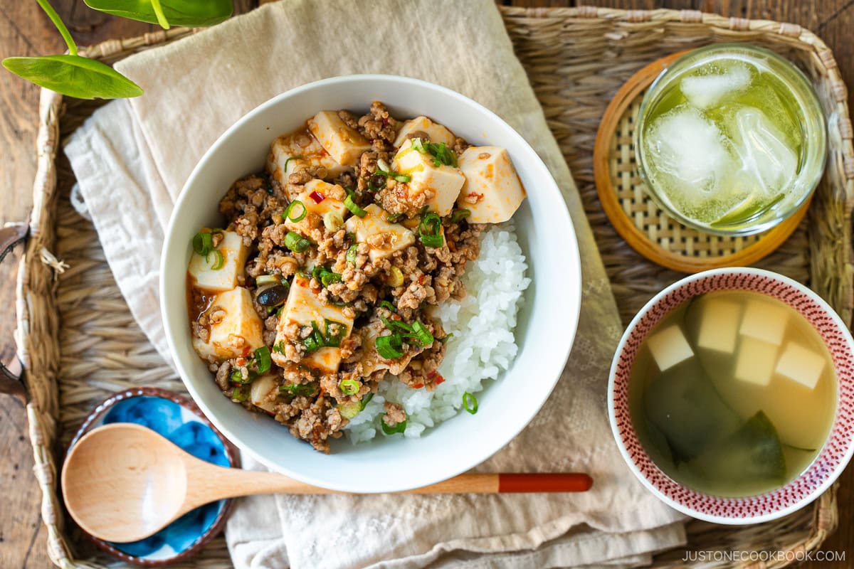 A donburi bowl containing mapo tofu over bed of steamed rice, served with a small bowl of miso soup and a glass of green tea.