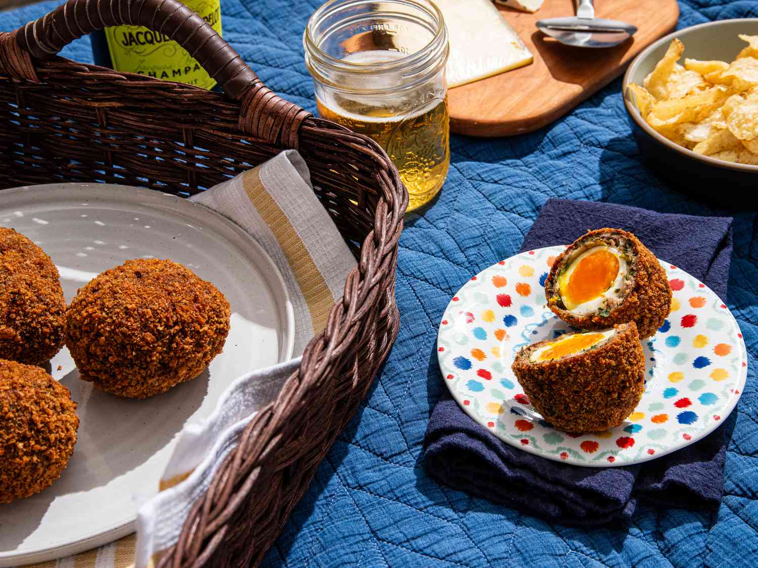 picnic basket with a plate of scotch eggs, and picnic set up with a plate of cut open scotch egg, chips, a drink, and a cheese wedge on a wooden board