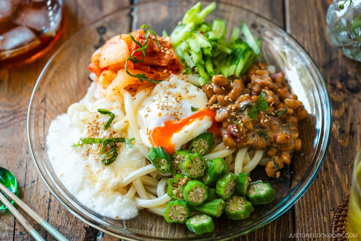 A glass plate containing cold natto udon topped with kimchi, onsen tamago, blanched okra, fresh cucumber, and grated nagaimo.