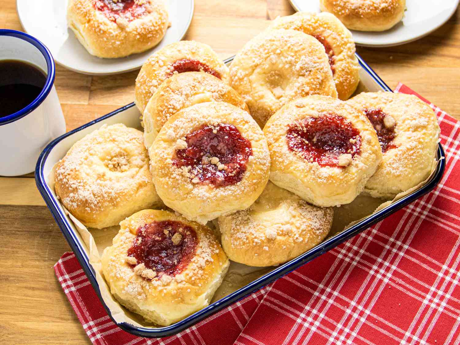 Kolaches on a platter, with one cream and one strawberry on 2 plates, with coffee on the side, red napkins, on a wooden surface. 