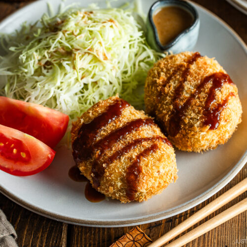 A round white plate containing Japanese croquettes, shredded cabbage, tomato wedges, and a tiny container with sesame dressing.
