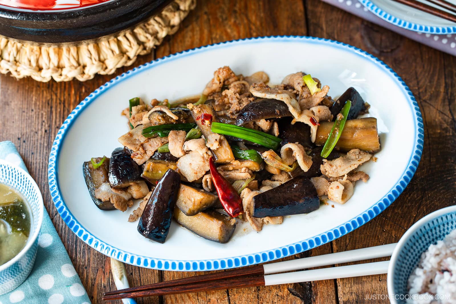 An oval plate with scalloped edge containing Pork and Eggplant Stir Fry served with multigrain rice and miso soup.