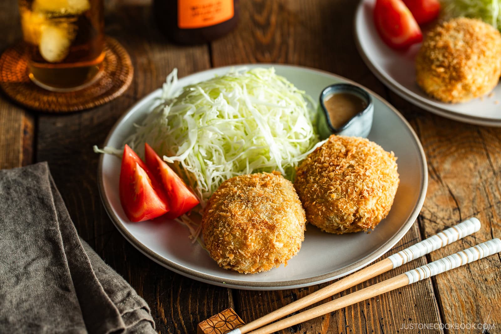 A round white plate containing Japanese croquettes, shredded cabbage, tomato wedges, and a tiny container with sesame dressing.