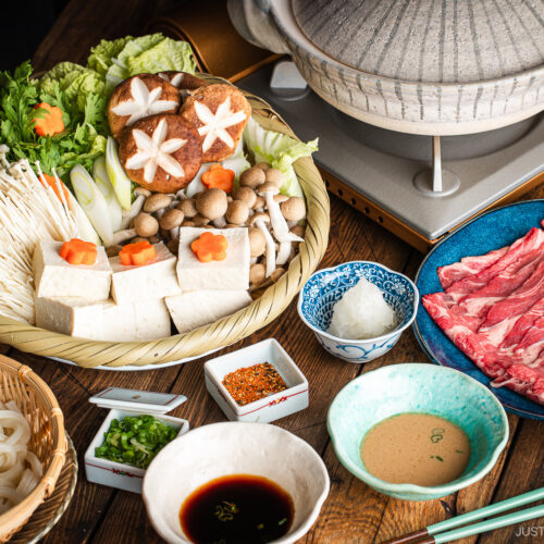 A Japanese shabu-shabu hot pot setup with a donabe pot, thinly sliced beef, tofu, assorted mushrooms, vegetables, udon noodles, and dipping sauces arranged on a wooden table.