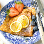 A blue plate containing lemon and sugar crepe along with fresh berries.
