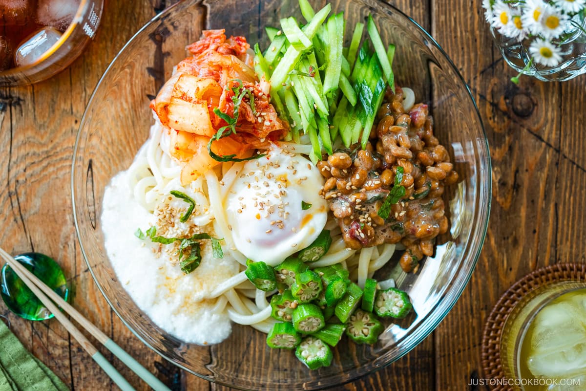 A glass plate containing cold natto udon topped with kimchi, onsen tamago, blanched okra, fresh cucumber, and grated nagaimo.