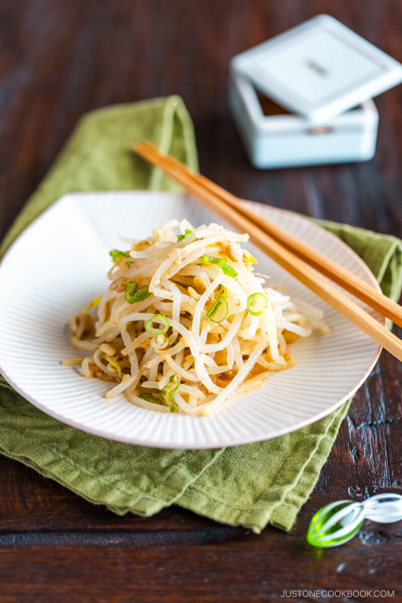 A white plate containing Bean Sprout Salad, seasoned with sesame oil and mixed with (ground) sesame seeds and green onions.