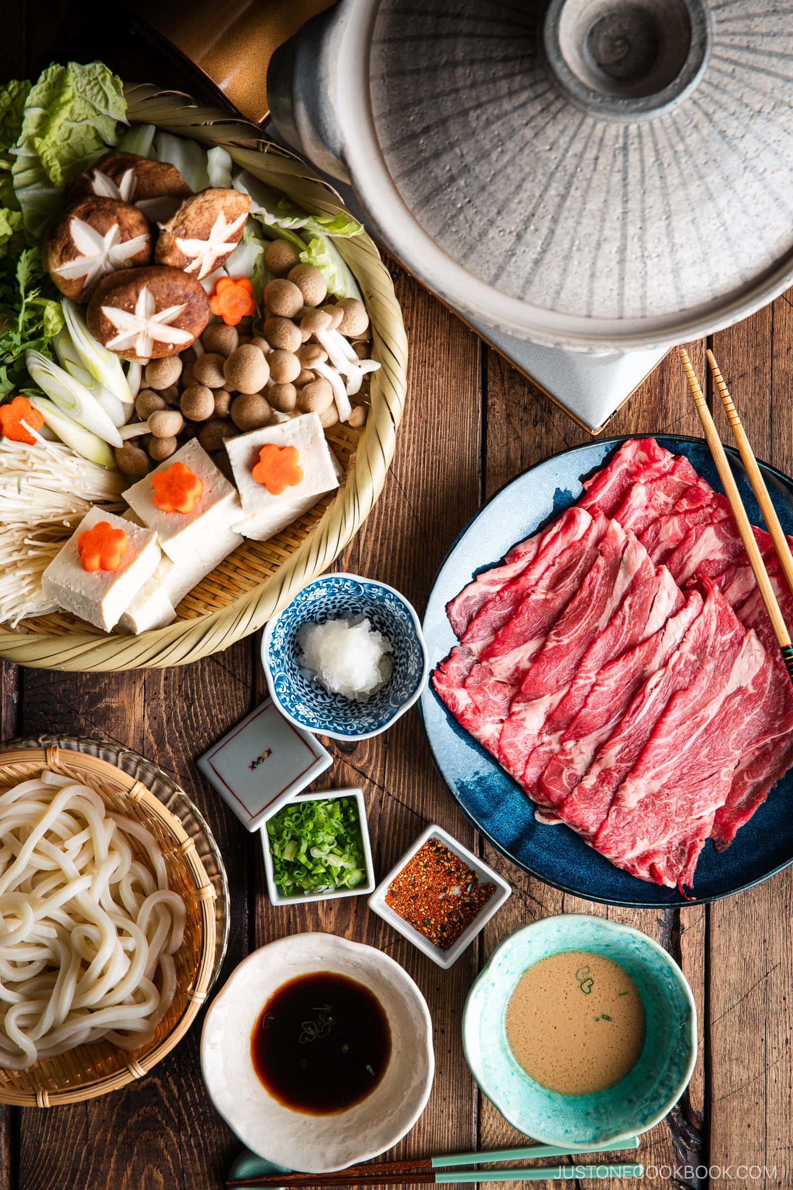 A Japanese shabu-shabu hot pot setup with a donabe pot, thinly sliced beef, tofu, assorted mushrooms, vegetables, udon noodles, and dipping sauces arranged on a wooden table.