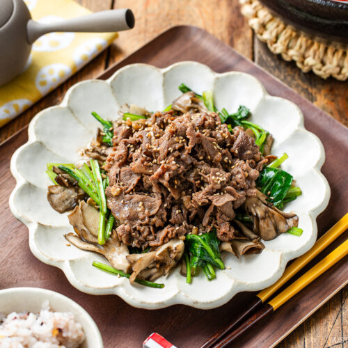 A white scalloped plate containing stir-fried Butter Ponzu beef with komatsuna green and maitake mushrooms.