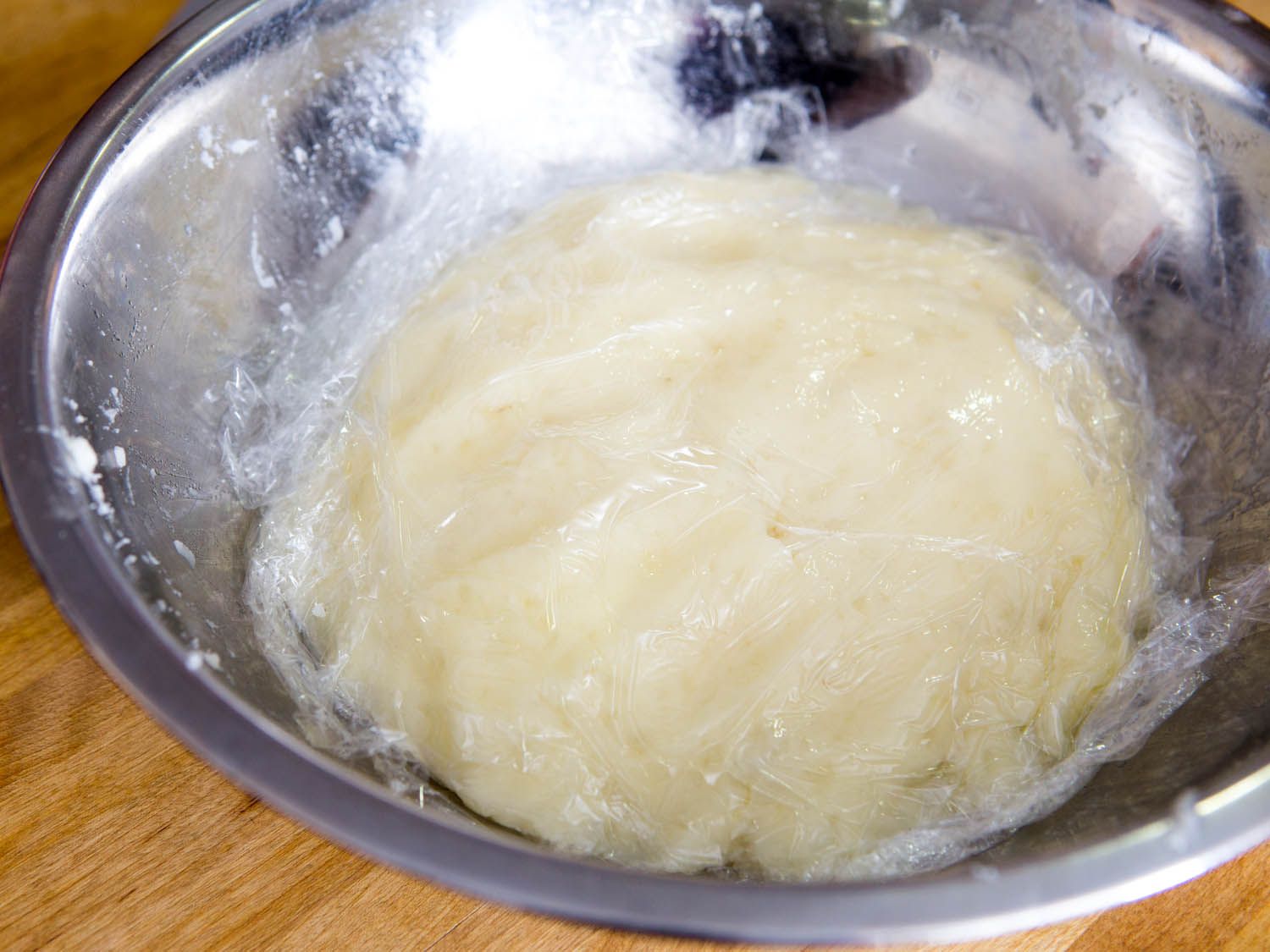 Mashed potatoes covered with plastic wrap in large silver bowl.