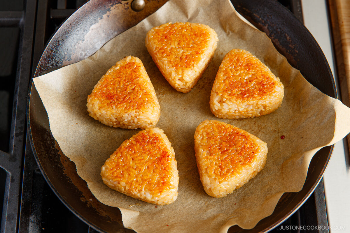 Five golden-brown grilled rice balls shaped into triangles are arranged on parchment paper in a frying pan.
