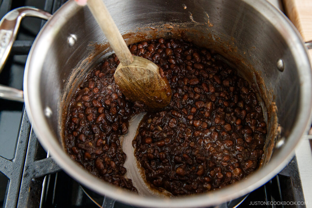 A pot of simmering sweetened red beans being stirred with a wooden spoon on a stove, with some beans pushed aside to reveal the thick liquid underneath.