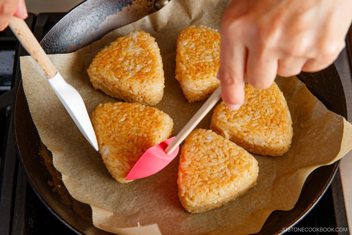Six golden-brown, triangular rice cakes are being cooked in a skillet lined with parchment paper. Two hands are turning a rice cake using a white and a pink silicone spatula.