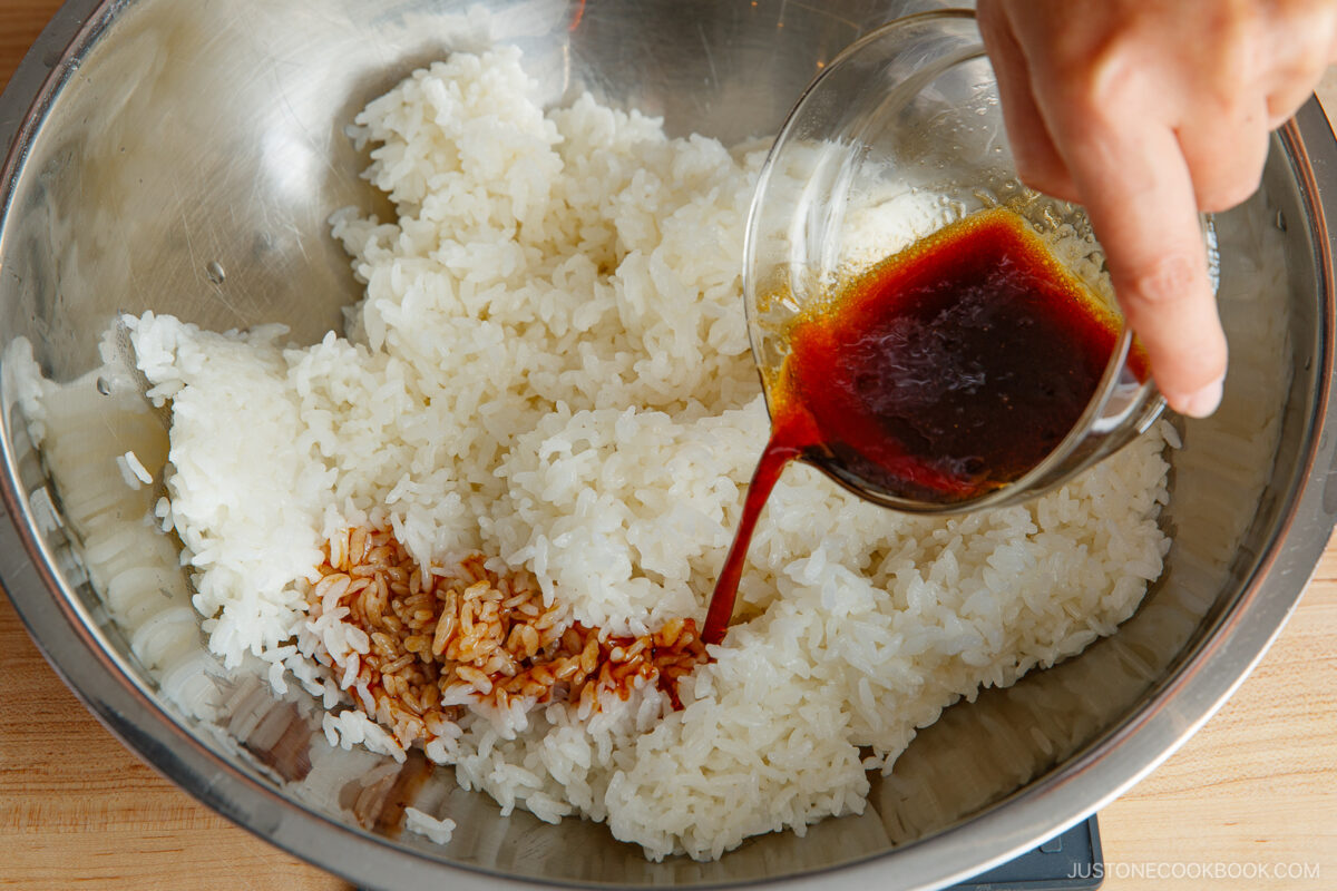 A hand pours a reddish-brown sauce from a glass cup onto white rice in a large metal mixing bowl on a wooden surface.