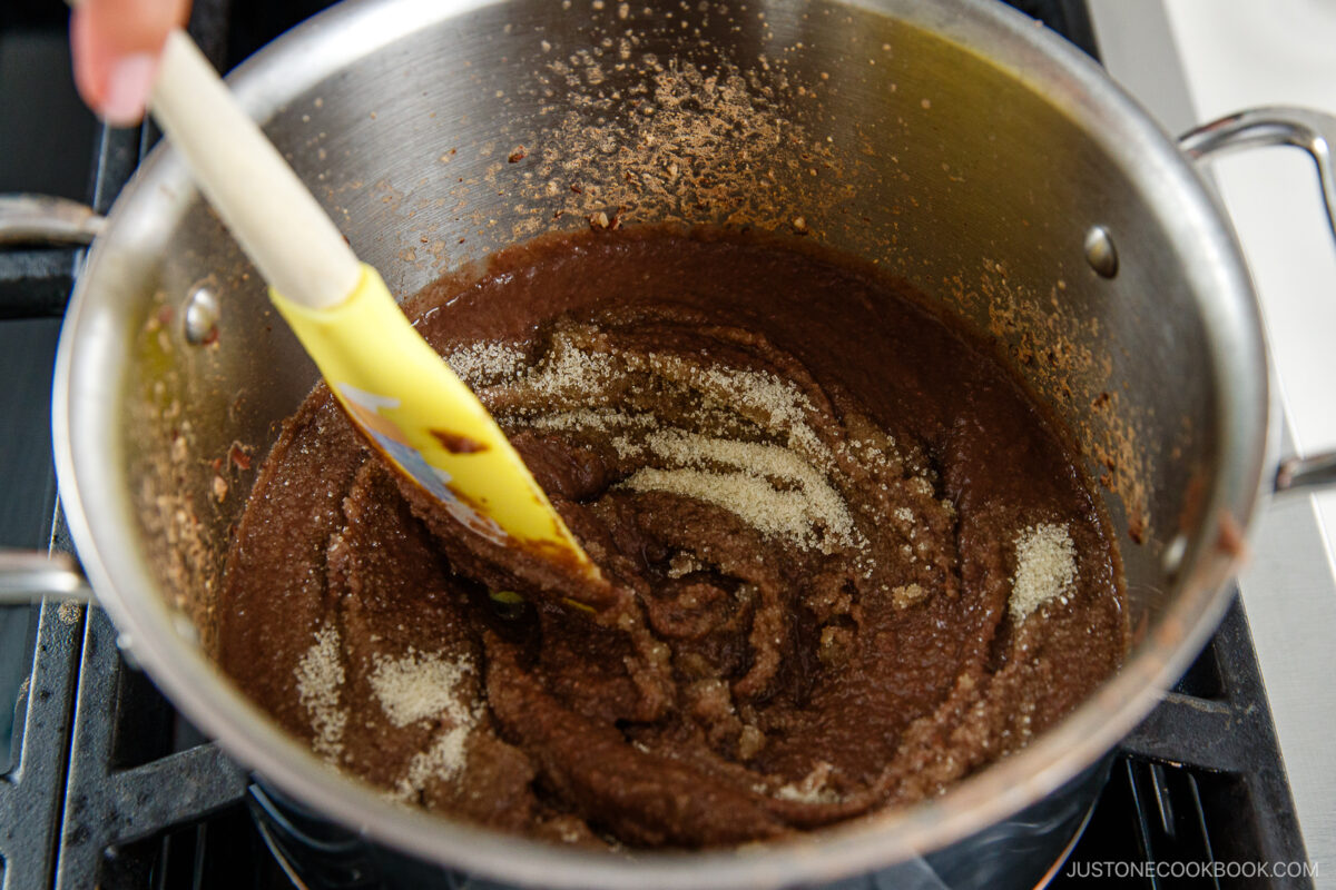 A hand stirs a thick brown mixture with a yellow spatula in a large metal pot on a stovetop, with some granulated sugar sprinkled on top of the mixture.