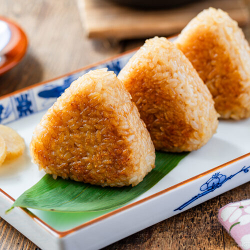 Three grilled Japanese rice balls (yaki onigiri) are arranged on a rectangular plate with a bamboo leaf, accompanied by pickled vegetables and a slice of pickled radish.