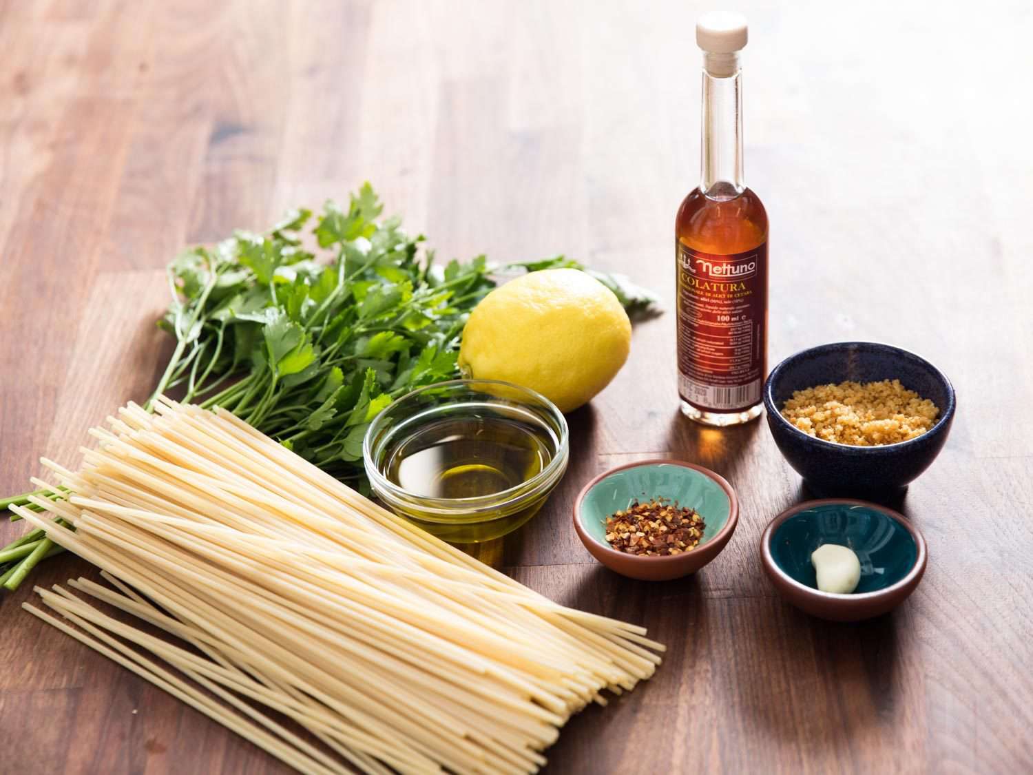 Ingredients for pasta con la colatura on a table: dried spaghetti, parsley, olive oil, lemon, chile flakes, bread crumbs, garlic, and a bottle of colatura di alici.