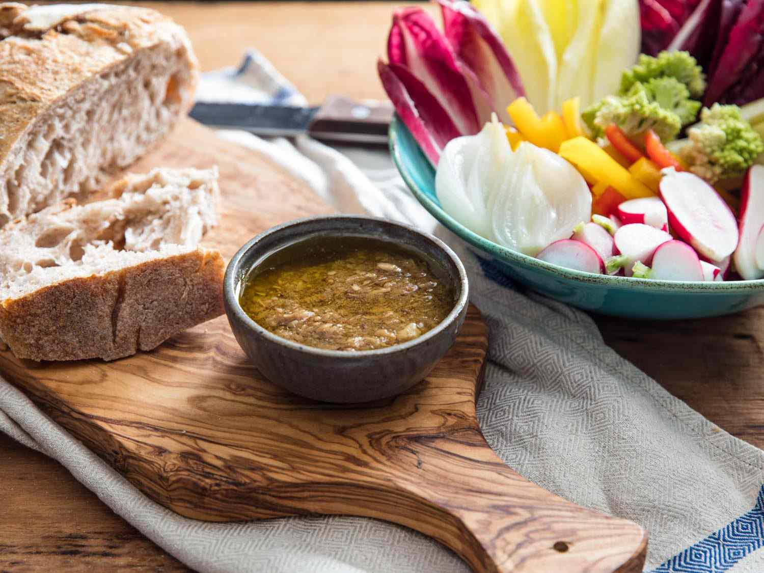 A bowl of bagna càuda dip on a wooden cutting board, with crusty bread and a bowl of colorful assorted vegetables nearby.