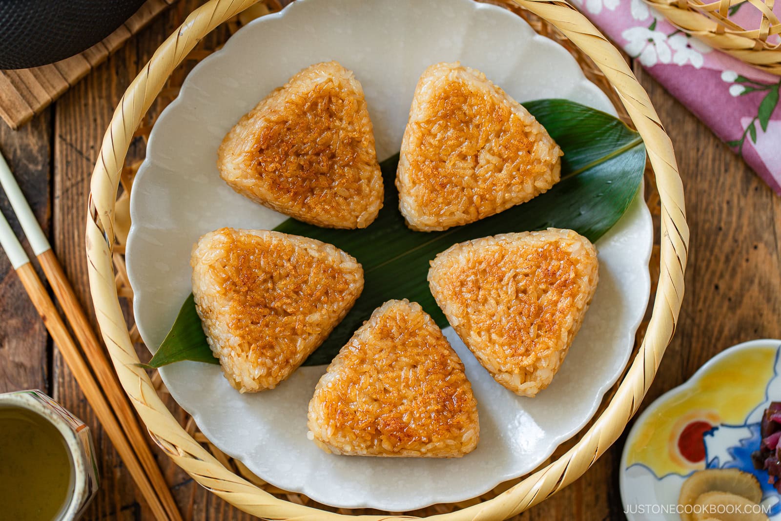 A plate with six golden-brown grilled rice balls arranged in a circle on a green leaf, set in a woven basket with chopsticks and small dishes nearby.