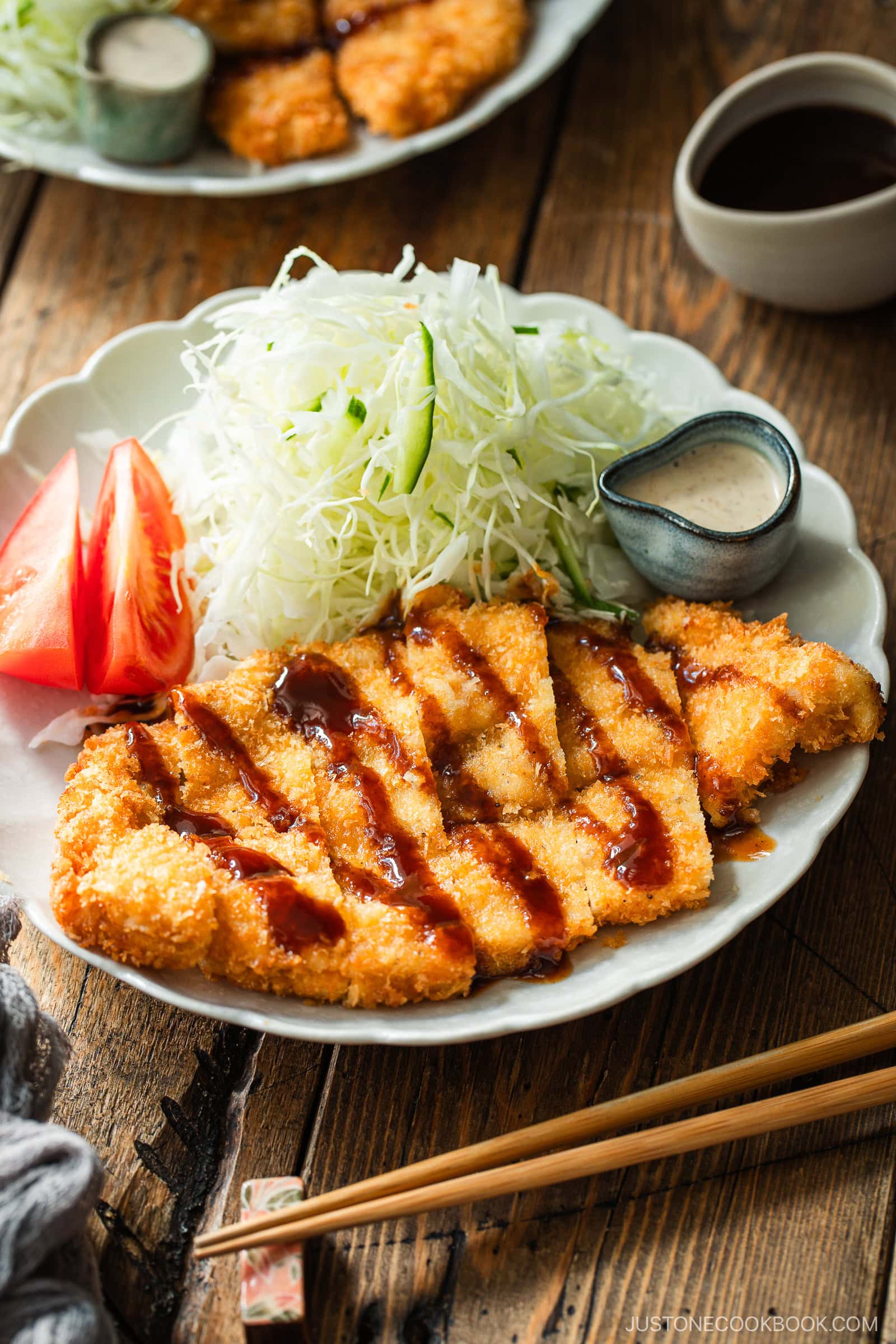 A plate of crispy breaded chicken cutlet with thick sauce, shredded cabbage, cucumber, tomato wedges, and a small dish of creamy dressing, served with chopsticks on a wooden table.