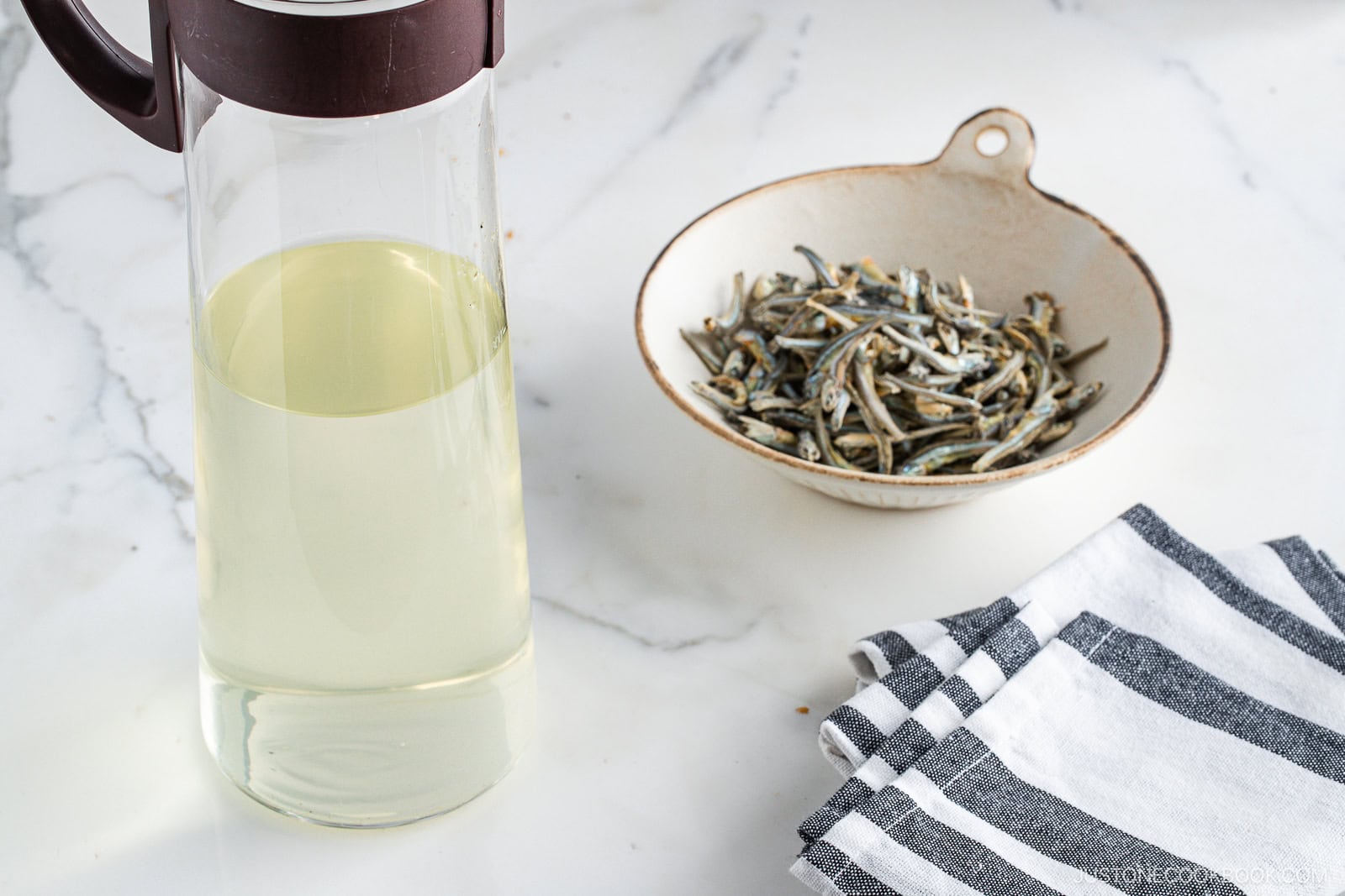 A glass pitcher filled with light yellow broth sits next to a bowl of dried anchovies and a folded striped kitchen towel on a white marble countertop.