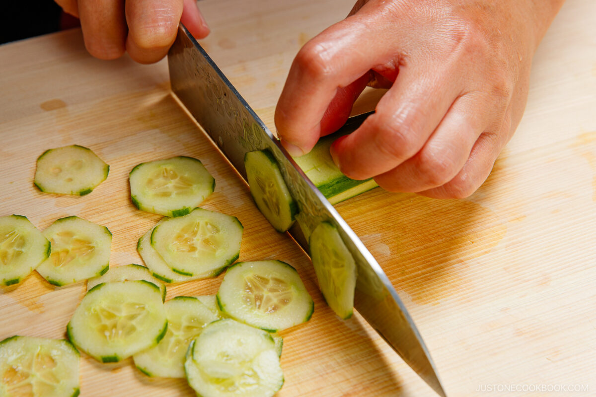 A person slices a cucumber into thin rounds on a wooden cutting board using a large kitchen knife.