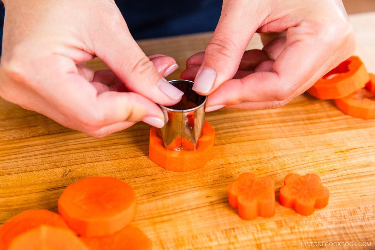 Close-up of hands using a small metal cutter to shape a sliced carrot into a flower on a wooden cutting board, with carrot slices and flower-shaped pieces nearby.