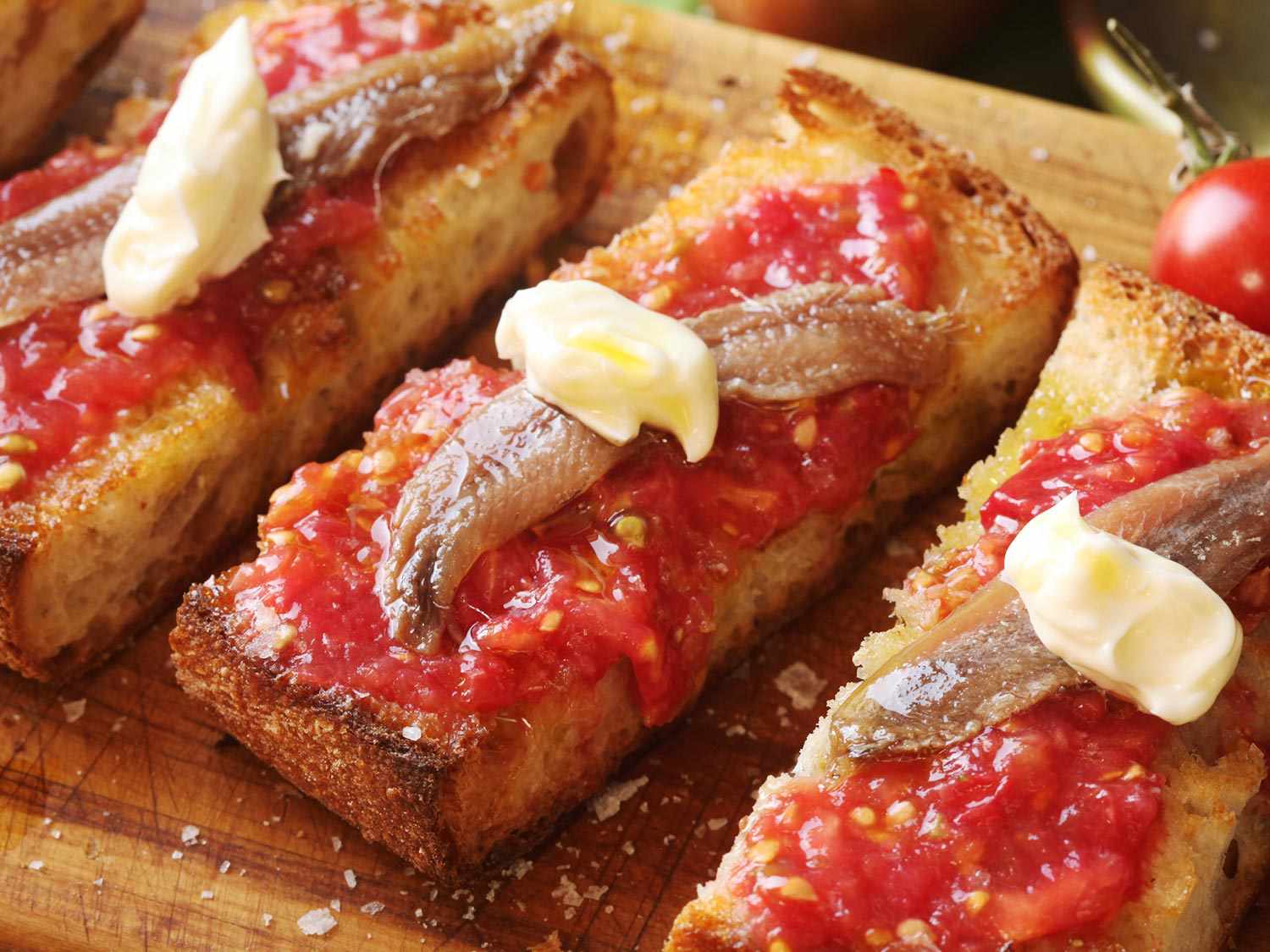 A close up of three slices of pan con tomate on a cutting board; each has an anchovy and a dollop of mayo on top. 