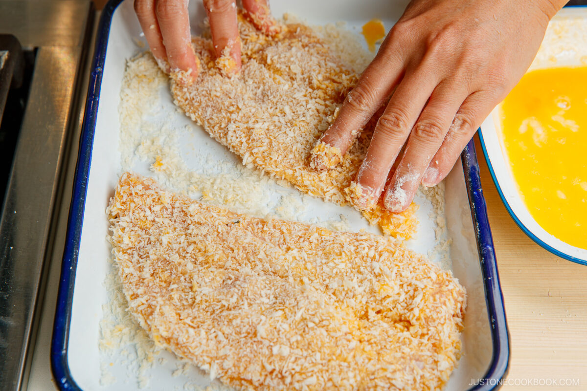 Two hands coat fish fillets in panko breadcrumbs on a white tray, with a bowl of beaten eggs nearby, preparing the fillets for frying.