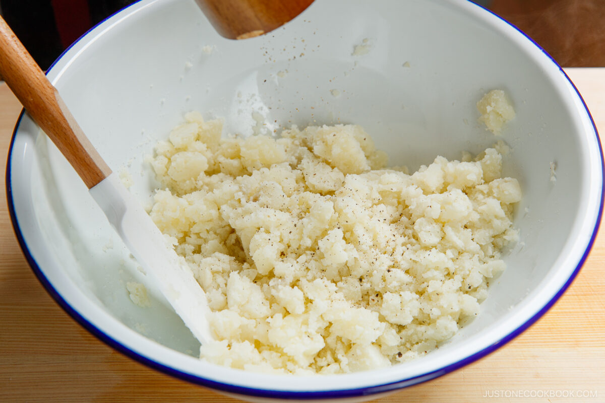 A white mixing bowl with japanese potato salad, black pepper sprinkled on top, and a spatula and wooden pestle resting inside the bowl.