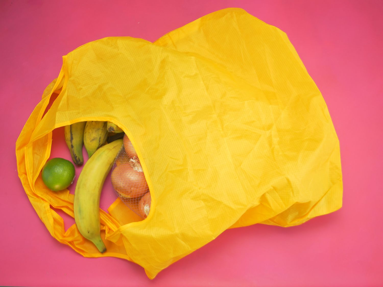 an orange Bee Green grocery bag on its side with plantains, onions, and limes spilling out