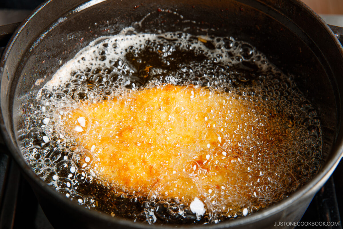 A breaded piece of food, likely fish or meat, is being deep-fried in hot oil in a black pan, with bubbles forming around the golden, crispy surface.