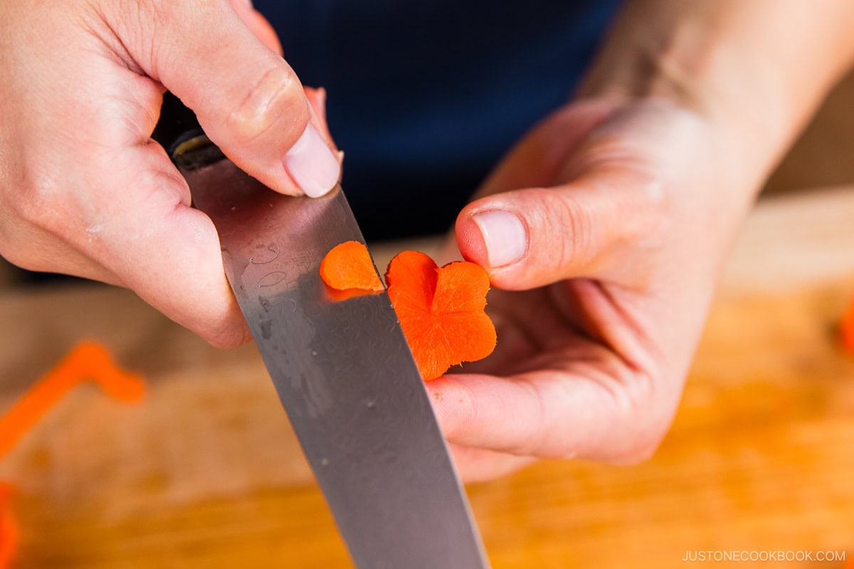 A person holds a knife and carefully carves a small orange carrot slice into a flower shape with their hands over a wooden cutting board.