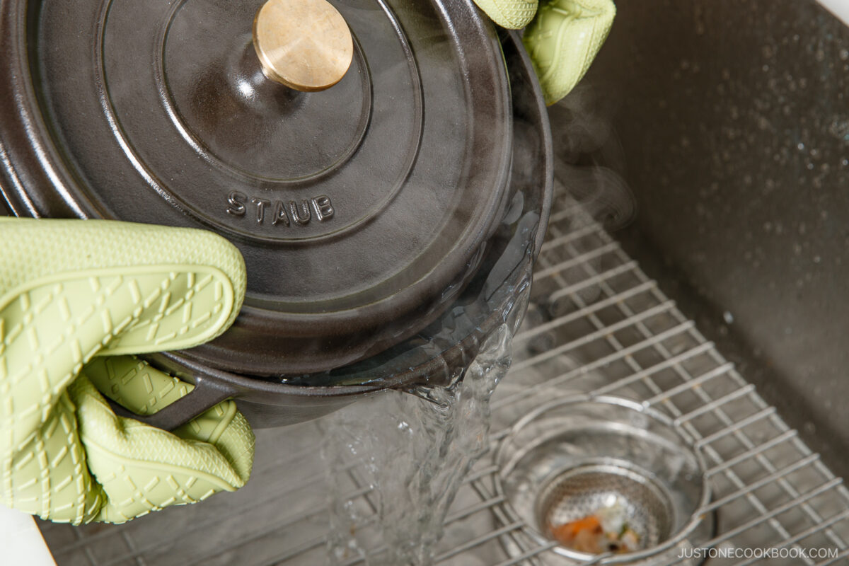 Wearing green oven mitts, a person pours hot water from a black Staub pot into a kitchen sink with a metal strainer—steam rises as they prep potatoes for japanese potato salad.