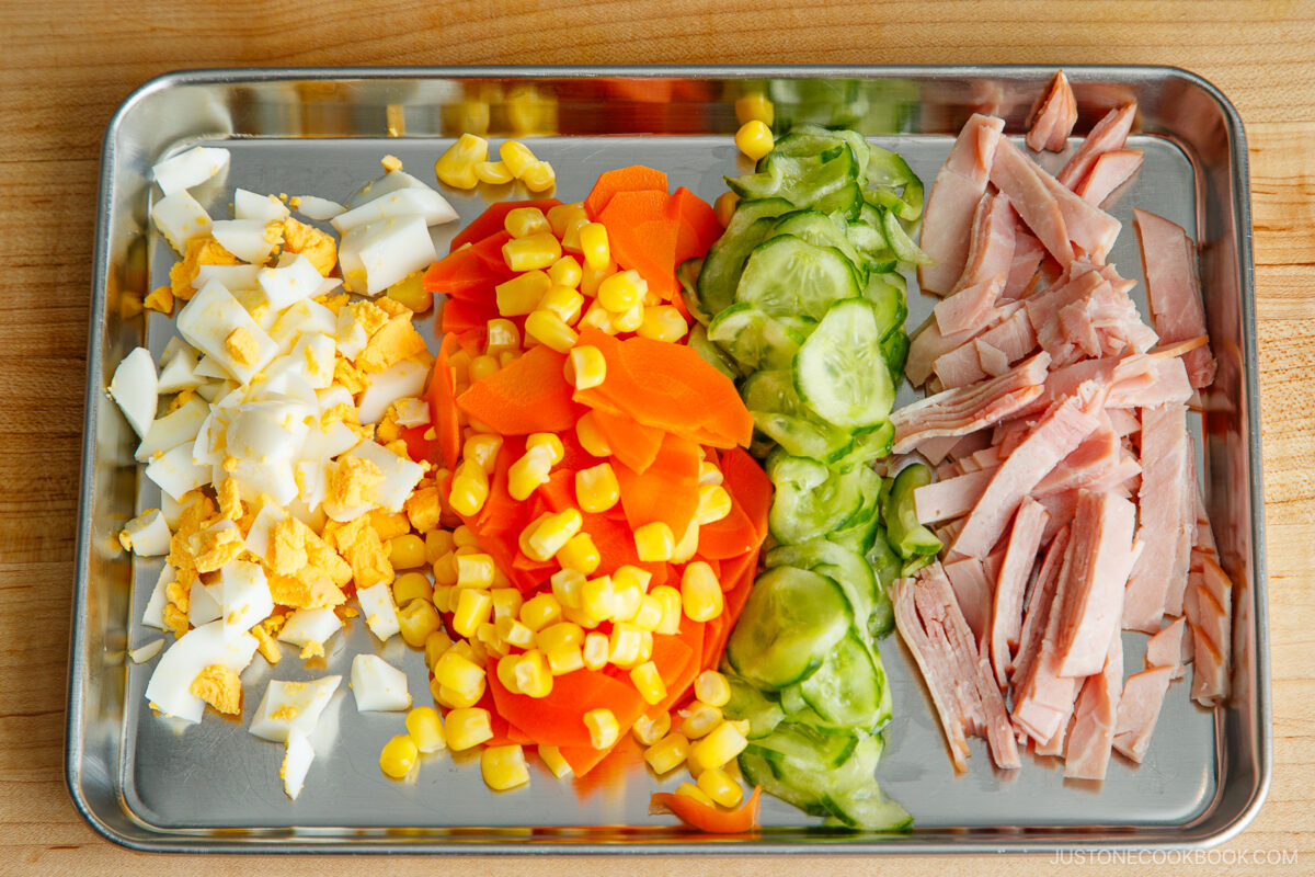 A metal tray with rows of chopped hard-boiled egg, corn kernels, sliced carrots, cucumber, and thinly sliced ham—perfect ingredients for making a classic Japanese potato salad—arranged neatly on a wooden surface.