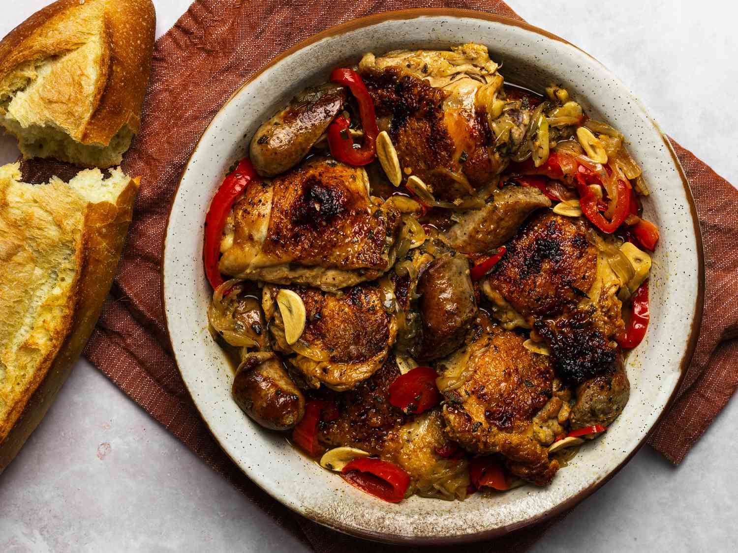 Chicken scarpariello in a round ceramic platter. The platter is on a maroon colored piece of cloth, and there is a broken loaf of bread on the left hand side of the image.