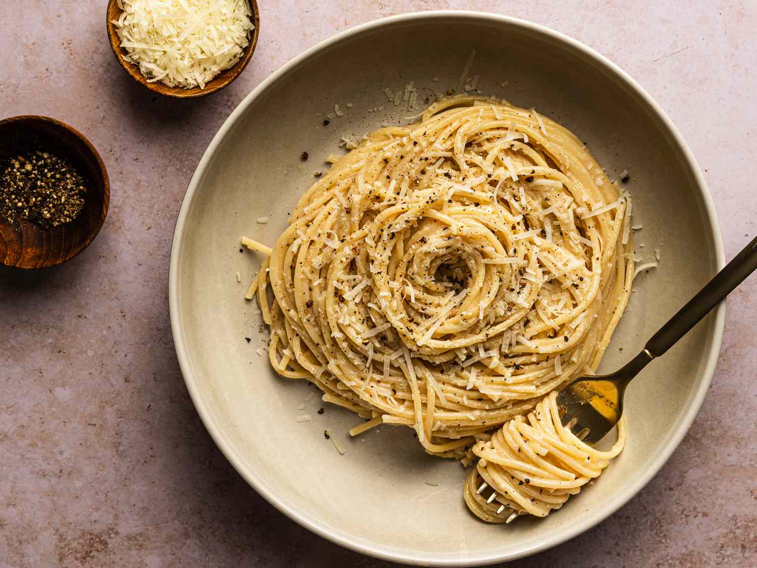 Cacio e pepe on a tan ceramic plate alongside two small bowls, one holding shredded cheese and the other holding freshly ground pepper. There is a metal fork with pasta swirled around it on the plate.