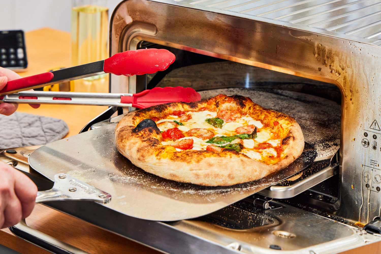 A pizza being removed from a countertop pizza oven with a steaming paddle and tongs