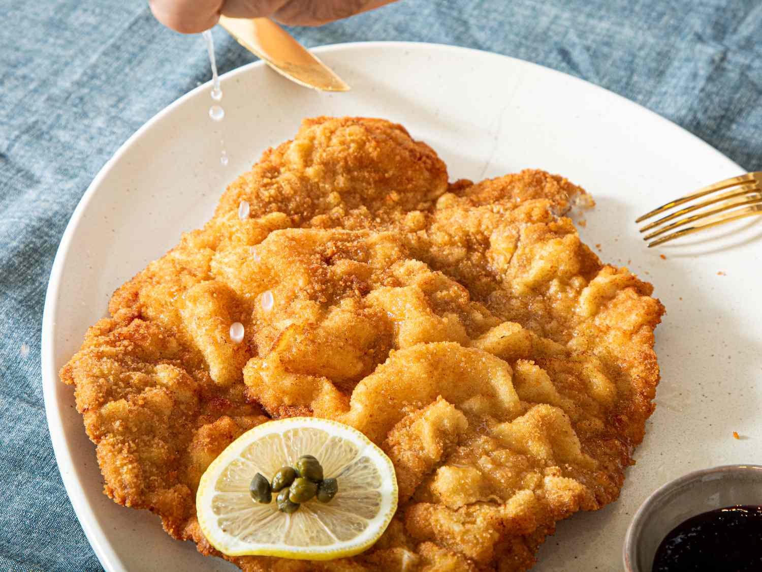 A plate of Wiener schnitzel on a table with a wedge of lemon and capers a hand squeezing lemon juice over it