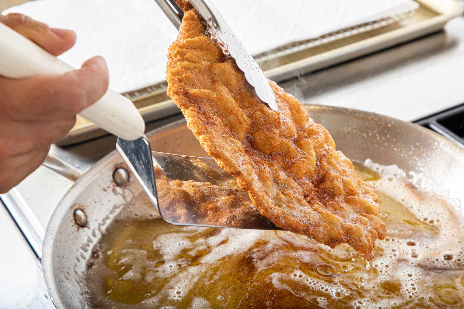 A breaded schnitzel being removed from a frying pan with tongs and a spatula