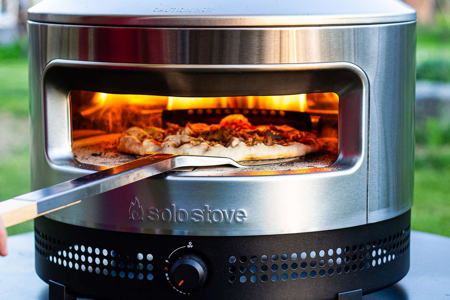 Pizza being cooked in an outdoor stainless steel Solo Stove Pi Prime, a peel positioned for retrieval