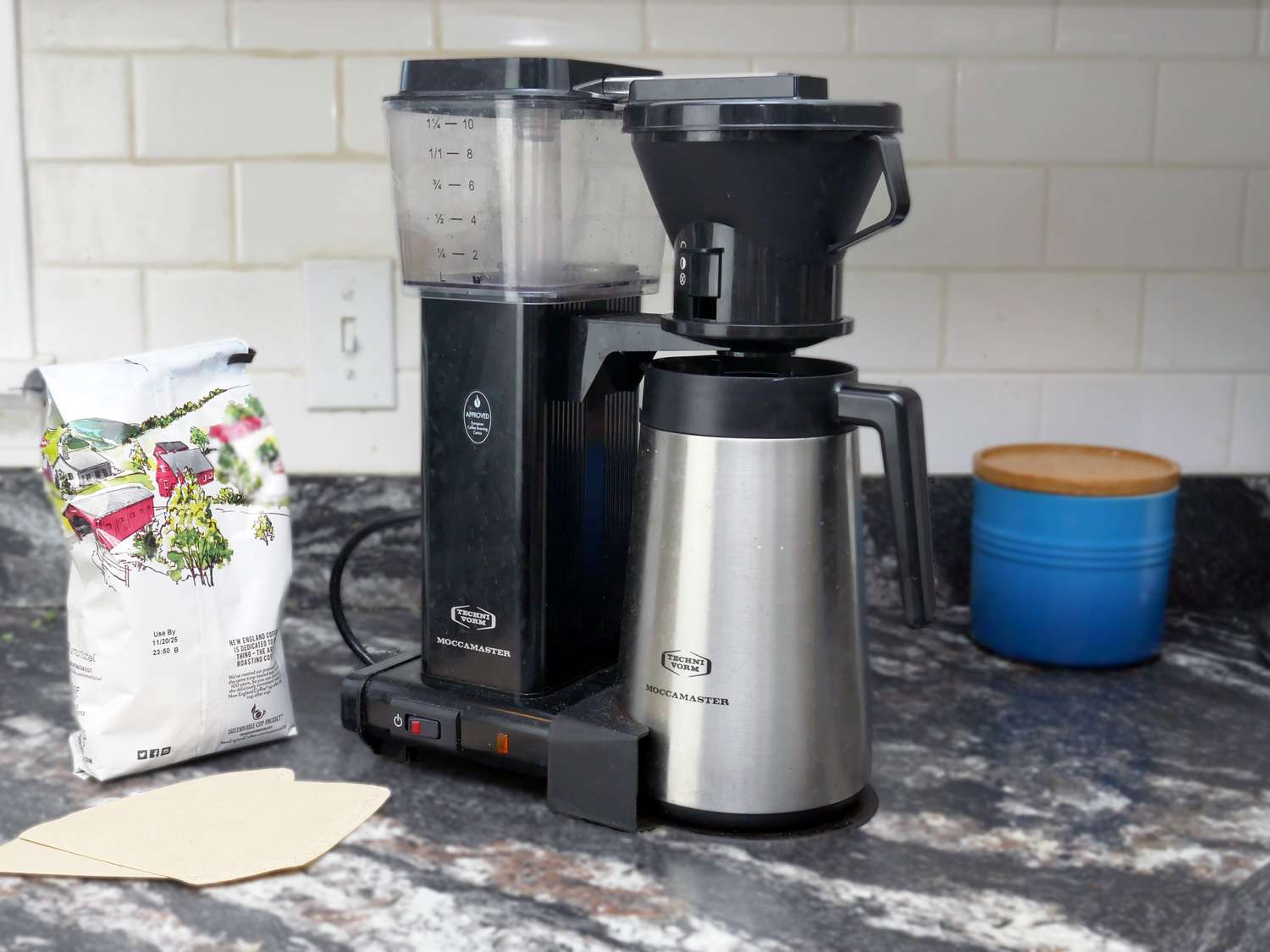 the technivorm moccamaster on a countertop with a bag of coffee, some filters, and a ceramic container of sugar. 
