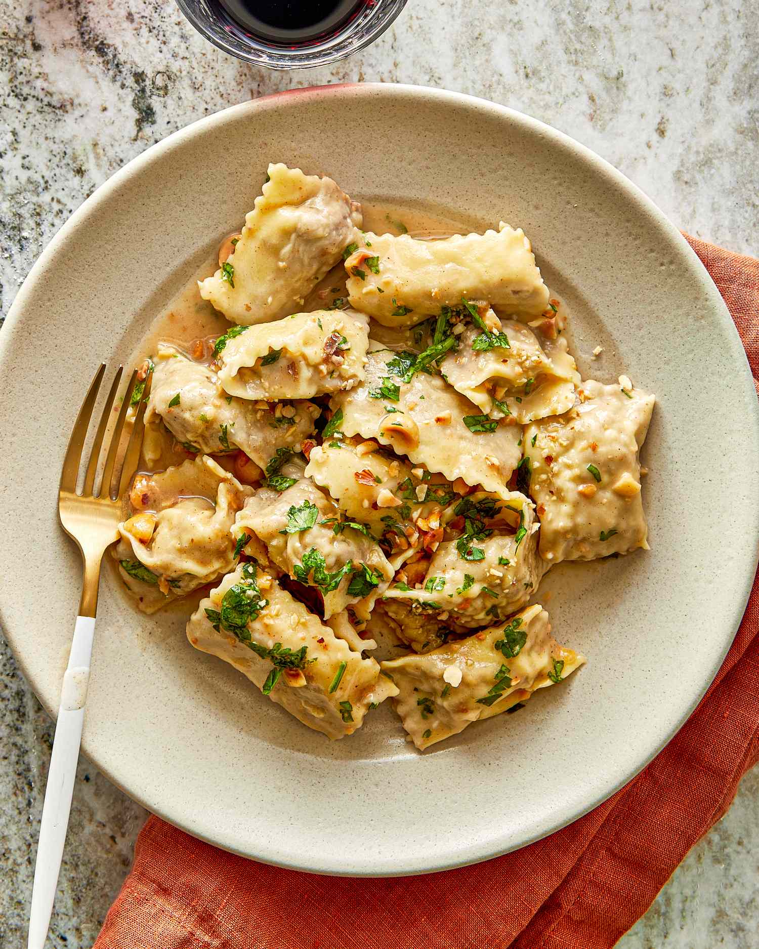 A plate of agnolotti pasta with garnishes served with a fork accompanied by a small glass of red wine