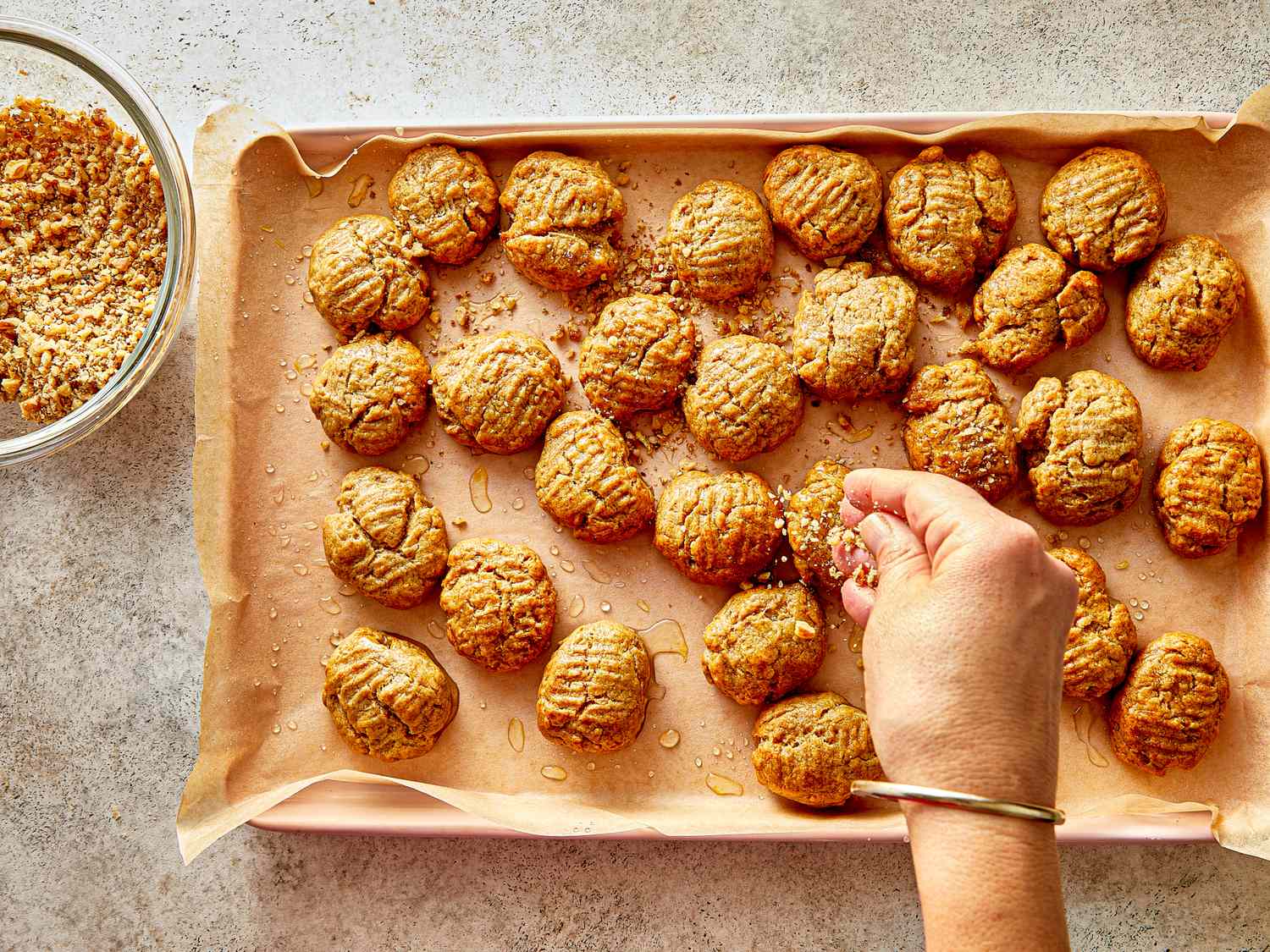 A persons hand sprinkling crumbs onto baked pastries on a tray lined with parchment paper with a bowl of crumbs beside them