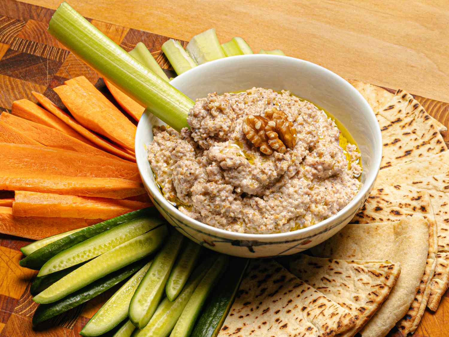 A bowl of walnut skordalia garnished with a walnut surrounded by sliced pita celery carrot and cucumber sticks